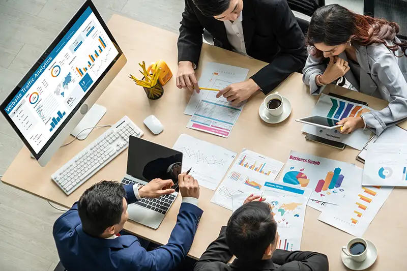 Team discussing analytics at desk with graphs and charts on monitor and paper Team discussing analytics at desk with graphs and charts on monitor and paper