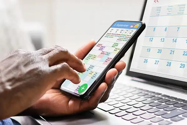 Hand holding a phone and scheduling an appointment on calendar, with laptop calendar in background